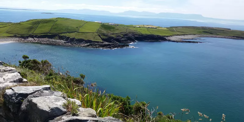 View of the seashore from stone wall. With headland in the foreground and mountains in the distance.