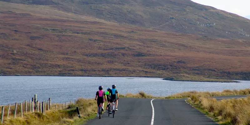3 cyclists wearing bright colours cycling towards a lake.