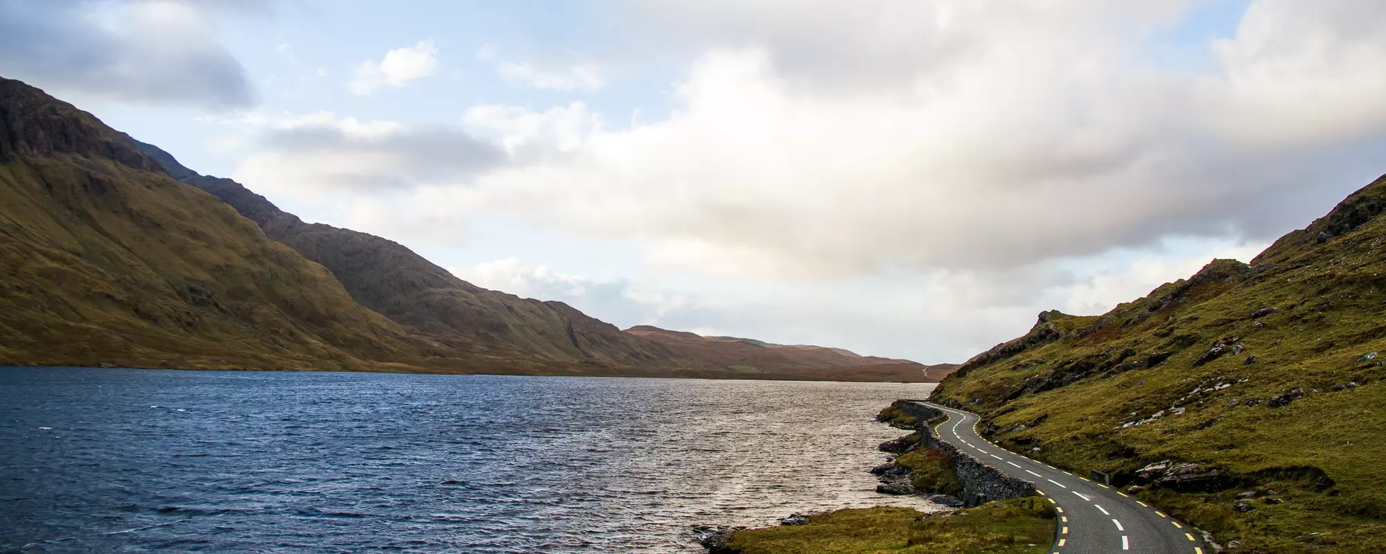 Lake with road running along side with hills each side under a partially cloudy sky.
