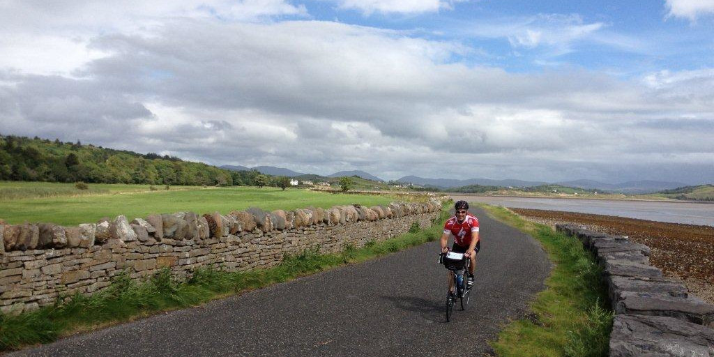 Cyclist wearing red cycling on quiet road with stone wall and green field.