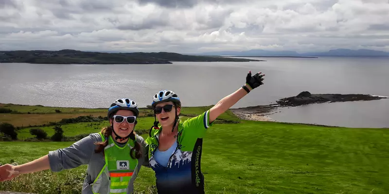 Two girls dressed in cycling gear waving in front of coast.