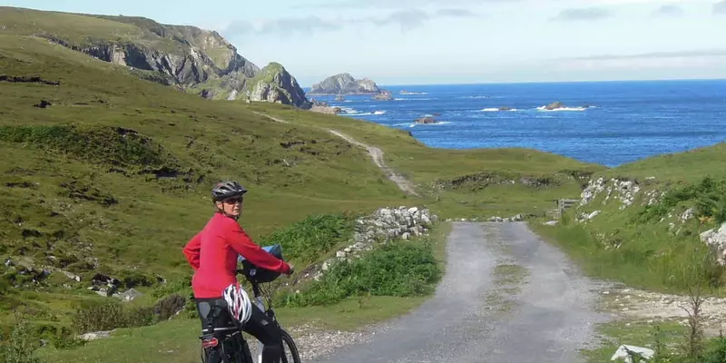 Cyclist wearing red standing on remote road leading to rocky shoreline